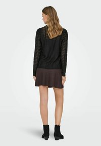 Svart strikket langermet genser med teksturert mønster, kombinert med et brunt skjørt og svarte ankelstøvler. Håret er bølgete og skulderlangt.