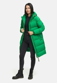 Femeie îmbrăcată cu o geacă lungă de puf verde strălucitor, un top negru, blugi skinny negri și cizme negre cu șireturi, stând pe un fundal alb.