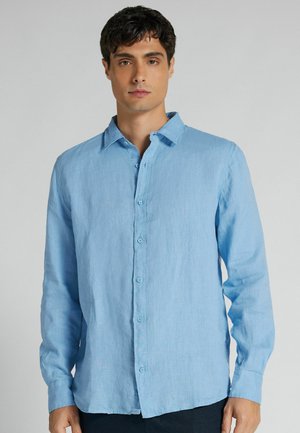 Mand iført en lyseblå skjorte med knapper og lange ærmer står foran en ensfarvet grå baggrund.