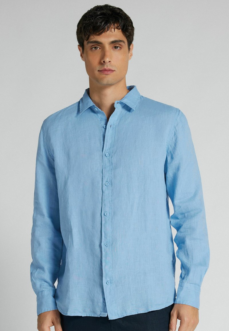 Homme portant une chemise bleu clair à manches longues boutonnée, debout devant un fond gris uni.