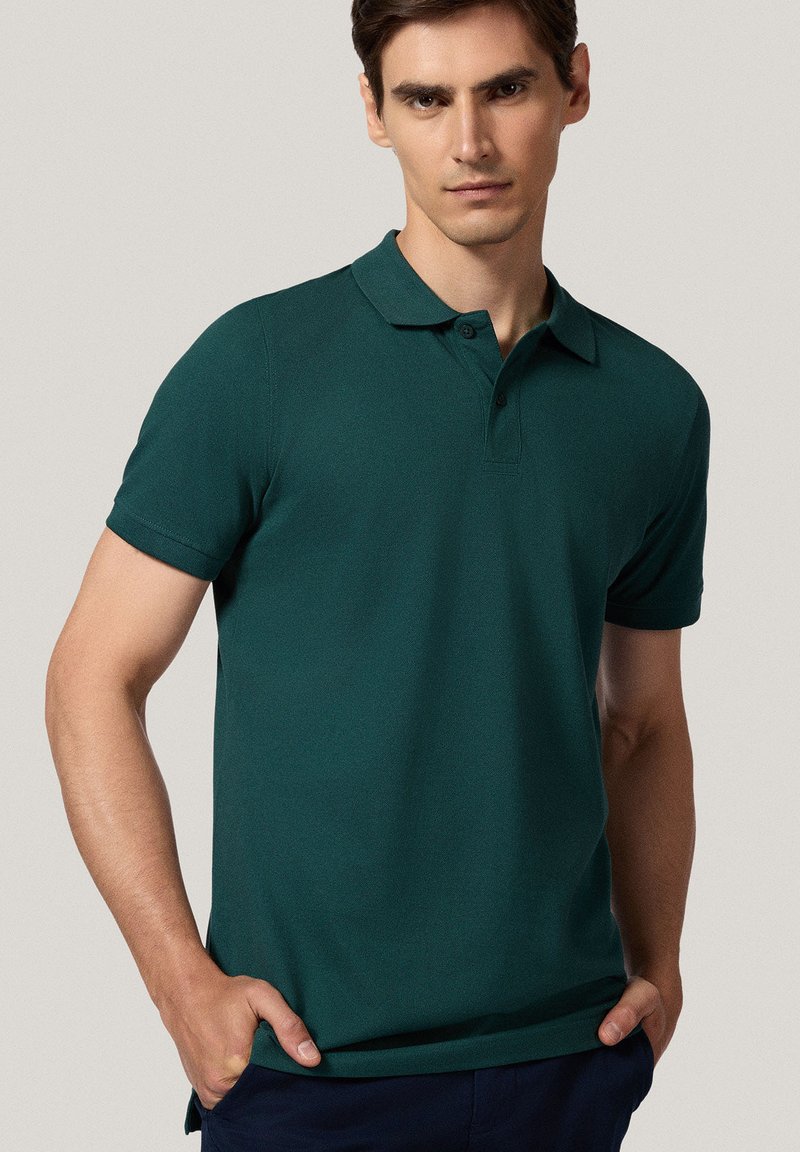 Ciemnozielona koszulka polo z krótkimi rękawami, płaskim kołnierzykiem, zapięciem na dwa guziki i miękką fakturą. Nosi w luźnym stylu, z dłońmi w kieszeniach.