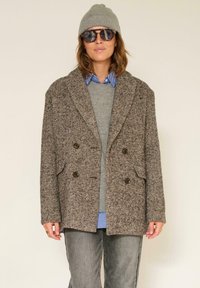Manteau croisé marron en chevrons avec grands revers, accents de boutons, assorti à un pull gris, une chemise rayée, un jean et un bonnet gris.
