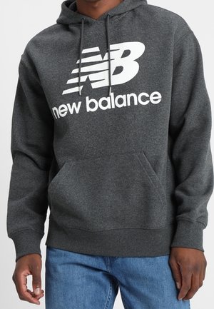 Ciemnoszara bluza New Balance z białym logo i napisem, noszona przez osobę w niebieskich dżinsach, stojącą na jednolitym tle.