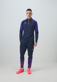Marineblauer Sportanzug mit lila Akzenten, bestehend aus einem Top mit halbem Reißverschluss und gemusterten, schmal zulaufenden Hosen. Knallpinke Schuhe runden den Look ab.