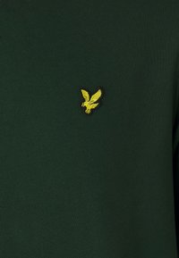 Sudadera verde de algodón con un pequeño logo bordado en amarillo de un águila en el lado izquierdo del pecho. Textura suave y cuello redondo clásico.