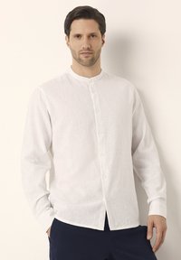 Camisa blanca de lino con manga larga y cuello mao, con frente abotonado y una textura sutil. Llevada con pantalones oscuros.