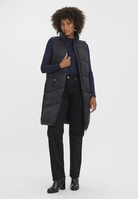 Svart vadderad lång väst med hög krage, dragkedjestängning och sidofickor, bärs över en marinblå långärmad tröja och svarta byxor. Glansiga stövlar.