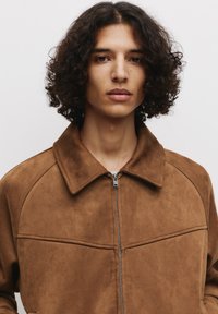 Veste en daim marron avec un col, dotée d'une fermeture à glissière devant et de détails en panneaux. Texture lisse et design minimaliste.