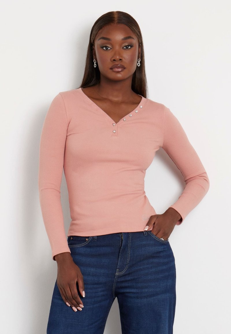 Rožinė su briaunomis ilgomis rankovėmis palaidinė su V formos iškirpte ir dekoratyviniais mygtukais vienoje pusėje. Suderinta su tamsiai mėlynomis džinsinėmis kelnėmis.