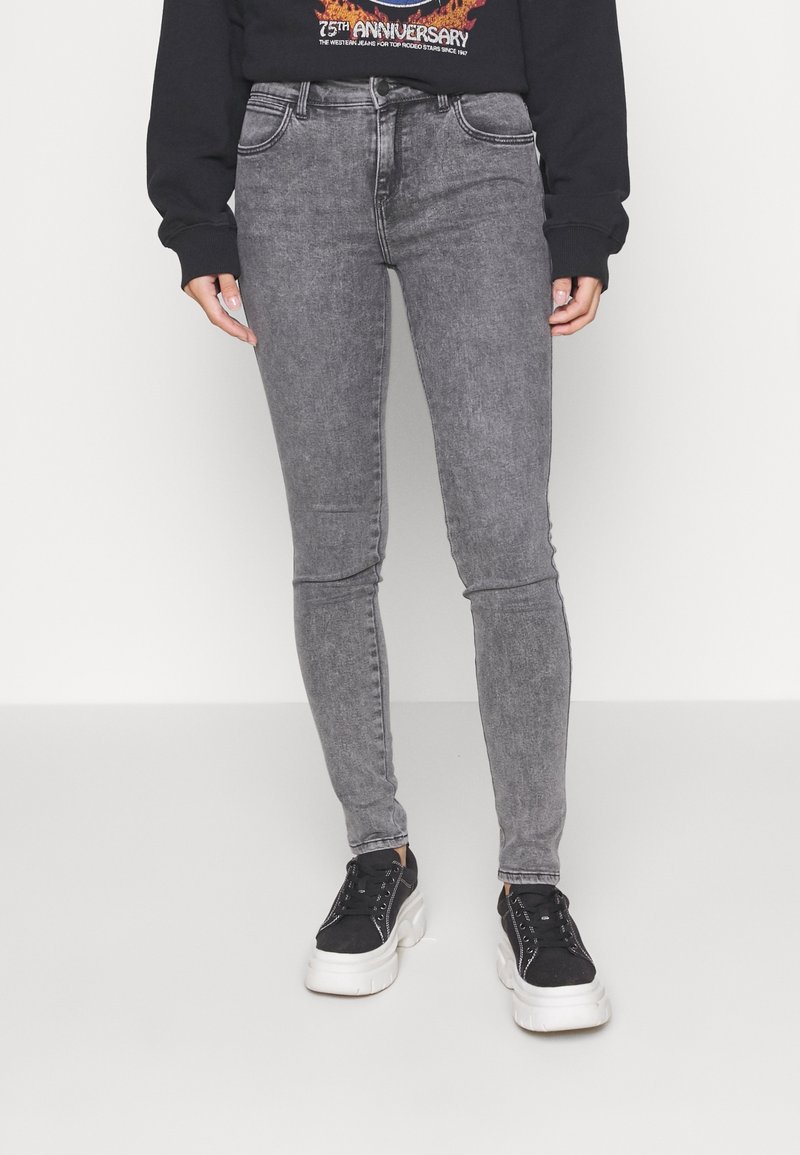 Jeans skinny de mezclilla gris con un acabado desgastado, cinco bolsillos y pasadores para cinturón, combinados con zapatillas negras y un sudadera gráfica negra.