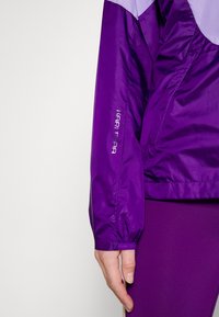 Veste légère violette avec une texture lisse, dotée d'un poignet élastique et d'un logo sur la manche. Poches latérales zippées visibles.
