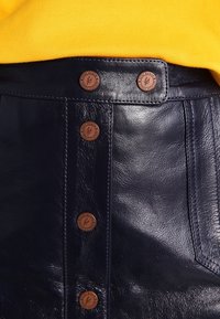Jupe en cuir marine avec cinq accents de boutons marron à l'avant, présentant des coutures tonales et une texture lisse. L'arrière-plan est un vêtement jaune.
