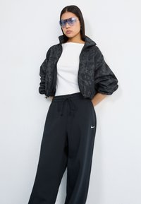 Una joven con el pelo liso lleva una chaqueta bomber negra con estampado, una camiseta blanca y pantalones de chándal negros, de pie con confianza contra una pared blanca.