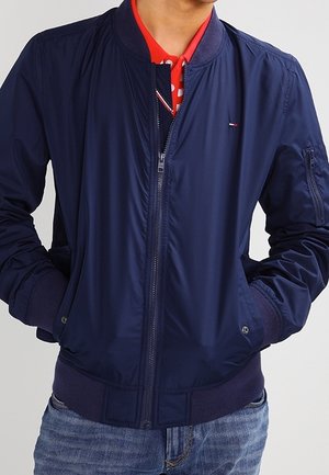 Hombre vestido con chaqueta bomber azul marino sobre una camisa polo roja y jeans azules, de pie con las manos en los bolsillos de la chaqueta.