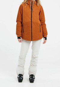 Braune gesteppte Jacke mit einer strukturierten Oberseite, schwarzem Reißverschluss, Seitentaschen und Kapuze, kombiniert mit hellen Skihosen und schwarzen Skischuhen.