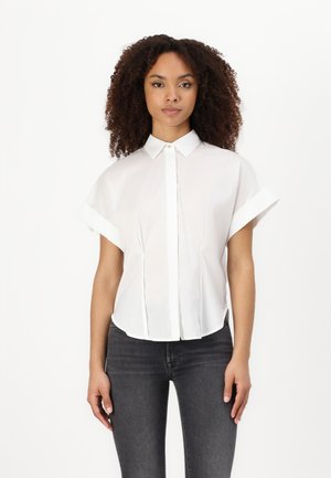 White short-sleeve blouse with a button-down front, curved hem, and folded cuffs, made from lightweight fabric. Worn with dark jeans.