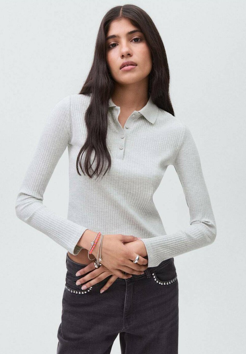 Ung kvinna med långt mörkt hår som bär en ljusgrå ribbad långärmad polo och mörka jeans med nitar på fickorna, händerna sammanflätade.