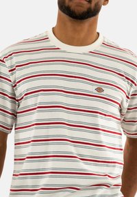 White short-sleeve shirt featuring horizontal red and blue stripes. Includes a small embroidered logo on the left chest and a ribbed neckline.