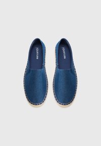 Chaussures slip-on en denim bleu avec des semelles tissées de couleur claire et des bords cousus. L'intérieur présente une doublure marine avec le nom de la marque en blanc.