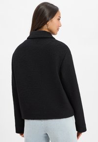 Black, textured jacket with a collared neckline, long sleeves, and a relaxed fit, shown from the back against a plain white background.