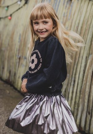 Zwarte sweatshirt met pofmouwen en bruine bloemenprint, gecombineerd met een geplooide metallic zilveren rok, tegen een textuur achtergrond.