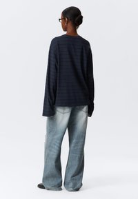 Μπλούζα σε σκούρο μπλε με λευκές ρίγες και μακριά μανίκια, με στρογγυλή λαιμόκοψη, συνδυασμένη με ανοιχτό μπλε τζιν παντελόνι με φαρδιά μπατζάκια, με ξεβαμμένη υφή και εμφανείς ραφές.