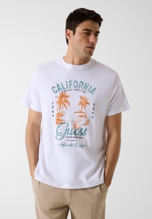 Man draagt een wit T-shirt met een oranjekleurige palmboomprint en tealkleurige tekst "California Guess Authentic Design", beige broek, handen in de zakken, kijkt naar rechts.