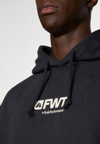Sudadera azul marina hecha de una tela suave con capucha ajustable. Presenta un logo blanco "FWT by PeakPerformance" en la parte frontal.