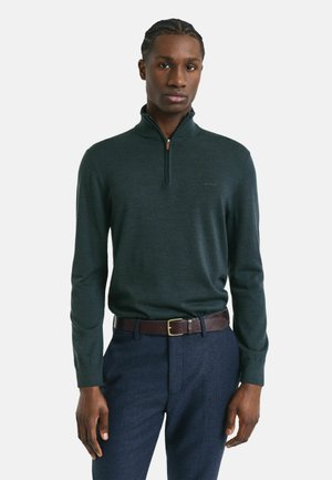 Donkergroene quarter-zip pullover van zacht materiaal, met lange mouwen en een subtiele logo op de borst, gecombineerd met marineblauwe broek en een bruine riem.