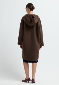 Brauner, mit Kapuze versehener Wollmantel mit weiten Ärmeln, knielangem Design und einer klaren, einfachen Silhouette. Getragen mit einem dunklen Jeansrock und burgunderfarbenen Schuhen.