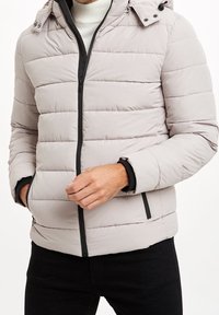 Světlounce šedá péřová bunda s horizontálním prošitím, černým zipem a nastavitelnou kapucí. Má boční kapsy a černé manžety s vnitřní podšívkou.