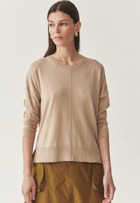 Beige kötött pulóver kerek nyakkivágással, es�ő vállakkal és bordázott ujjakban. Puha textúra, laza szabás, középen varrott részlettel.
