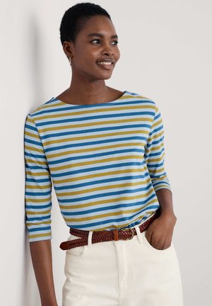 Gestreiftes Langarmshirt in Blau, Gelb und Weiß mit einem Bootsausschnitt. Kombiniert mit hellfarbigen Hosen und einem braunen geflochtenen Gürtel.