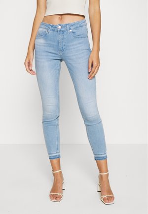 Frau trägt hellblaue Skinny-Jeans, die oberhalb der Knöchel gekürzt sind, weiße Sandalen mit Blockabsatz und Riemchen sowie ein kurzes weißes Oberteil.