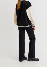 Černá a krémová varsity bunda volného střihu s kontrastními rukávy a žebrovanými manžetami, v kombinaci s černými rozšířenými džínami a robustními botami.