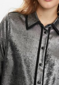 Camicia grigia metallica con una texture lucida, colletto nero e pattina dei bottoni nera. Presenta una superficie liscia e cinque bottoni neri.