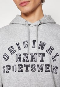 Gros plan d'une personne portant un sweat à capuche gris clair avec l'inscription « ORIGINAL GANT SPORTSWEAR » imprimée en grandes lettres noires sur la poitrine.