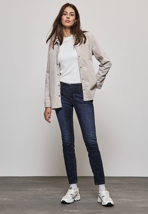 Chemise boutonnée gris clair sur un t-shirt blanc, associée à un jean bleu foncé et des baskets blanches. Textures lisses avec des motifs subtils.