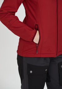 Röd mjuk skaljacka med dragkedjefickor och elastiska mudd, med en slät yta och logotypdetalj på ärmen.