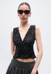 Μαύρο τοπ με παγιέτες, βαθύ V-λαιμόκοψη και φαρδιές τιράντες· υφή με εφαρμοστή γραμμή. Φορεμένο με γκρι ριγέ φούστα και σκούρα γυαλιά ηλίου.