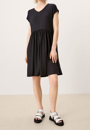 Mujer que lleva un vestido negro suelto de manga corta con escote en V y cintura fruncida, combinado con sandalias blancas de plataforma con tiras.