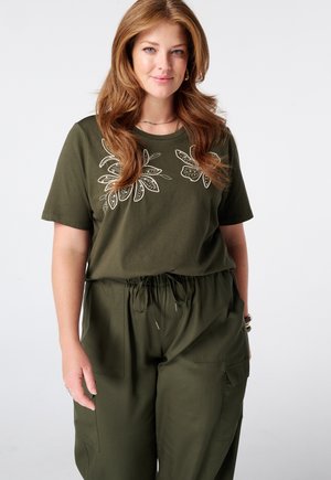 Femme aux longs cheveux châtain clair portant un t-shirt vert olive brodé de fleurs et un pantalon à cordon assorti, debout devant un fond uni.