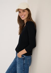 Lächelnde Frau, die eine beigefarbene Mütze, ein schwarzes Langarmshirt und blaue Jeans trägt, lehnt mit den Händen in den Taschen gegen eine beigefarbene Wand.