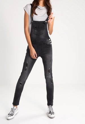 Sorte denim overalls med en tætsiddende silhuet, slidte detaljer, justerbare skulderremme og sølvfarvede knapdetaljer. Båret med en grå top og sølvfarvede sneakers.