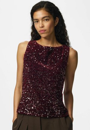 Femme aux cheveux bouclés et foncés, portant un haut sans manches à sequins rouge foncé et un pantalon marron foncé sur un fond clair uni.