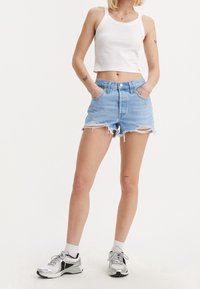 Shorts de mezclilla azul claro con dobladillo deshilachado, de tiro alto y diseño de cinco bolsillos. Combinados con una camiseta tipo cropped blanca y zapatillas deportivas plateadas.