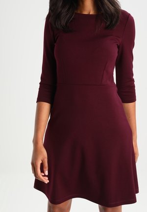 Femme portant une robe bordeaux ajustée, longueur genou, avec des manches trois-quarts, sur fond blanc.
