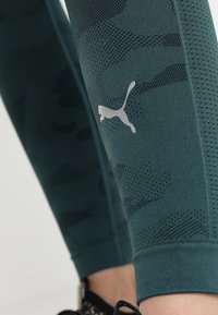 Zbliżenie na ciemnozielone, teksturowane legginsy z odblaskowym logo Pumy nad kostką, w połączeniu z czarnymi butami sportowymi.