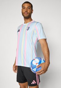 Gestreiftes hellblaues, pinkes und gelbes Trikot mit Rundhalsausschnitt und Juventus-Logo. Modell hält einen blau-weißen Fußball. Schwarze Shorts mit pinken Streifen.