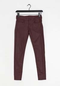 Burgundské skinny džíny s hladkou texturou, předními kapsami a zapínáním na zip, vystavené na černém závěsu na bílém pozadí.